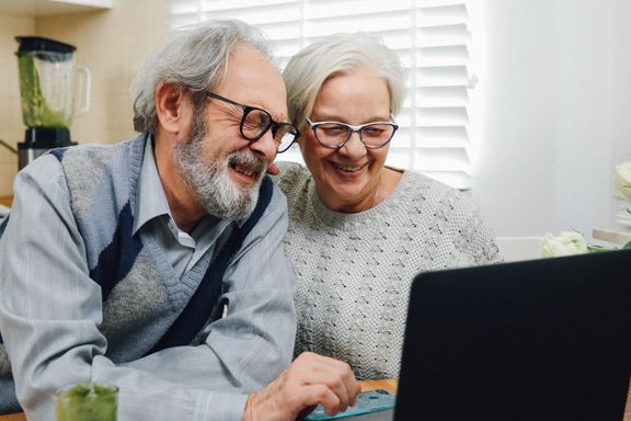 An older couple leans in close and laughs together at a laptop in their kitchen