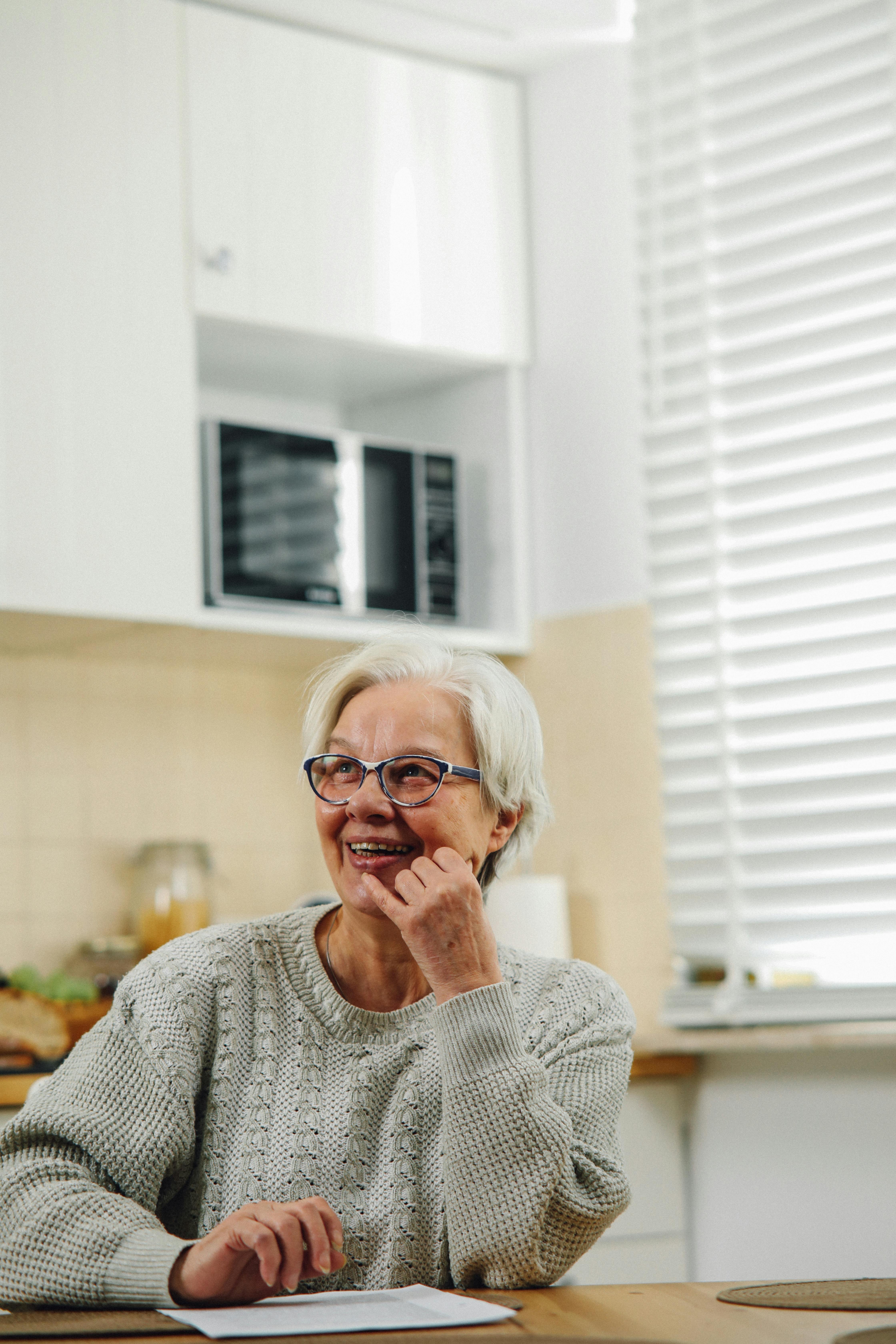 A cheerful elderly woman with glasses smiles warmly as she sits in a bright kitchen, adding a sense of comfort and homeliness.