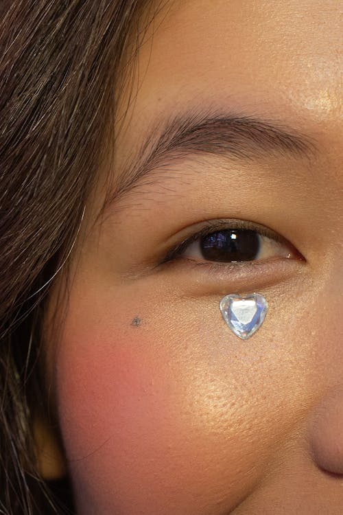 Crop positive Asian female with shiny crystal on skin under eye glancing at camera contentedly