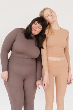Positive young plus size and fit female models with dark and blond hair in activewear holding hands and cuddling with closed eyes while standing in white studio