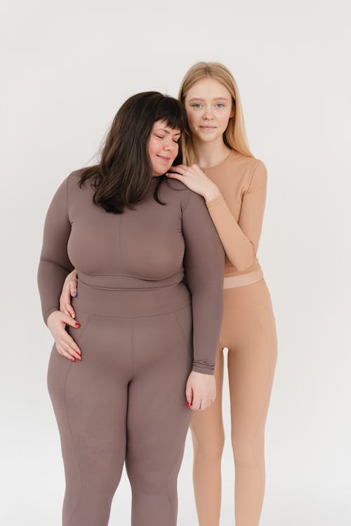 Positive young slim lady with blond hair in activewear smiling and embracing obese female friend while standing together against white background in studio