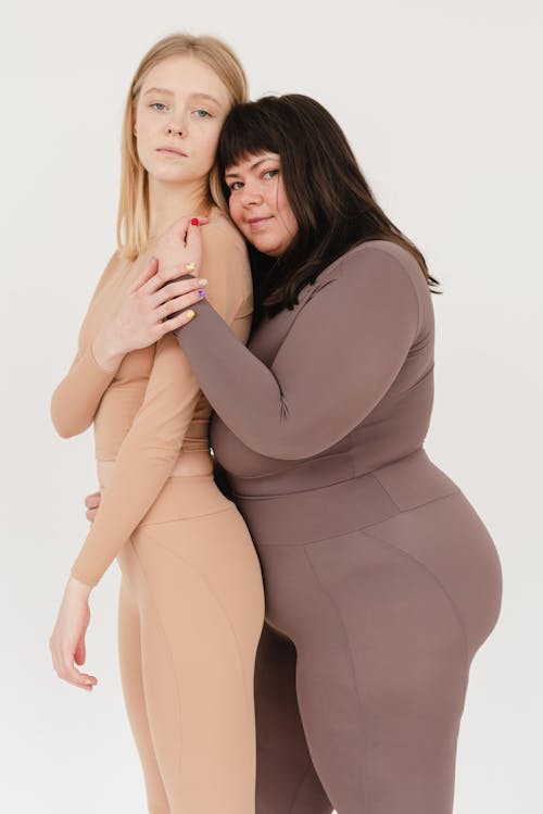Confident young curvy and fit ladies hugging and looking at camera against white background