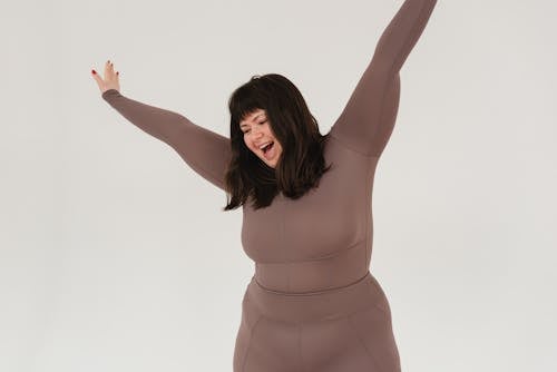 Cheerful young plus size female model with dark hair in activewear yelling happily while standing in white studio with raised arms