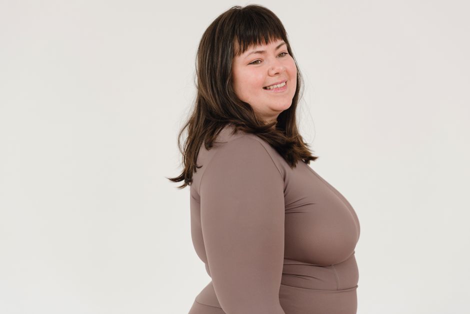 Side view of happy young overweight female model with long dark hair in activewear smiling and looking away against white background
