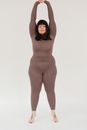 Full length of overweight young barefooted lady with dark hair in sportswear stretching body with raised arms and looking at camera before yoga practice against white background