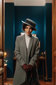 Stylish woman in a gray coat and hat, standing indoors with vintage decor.