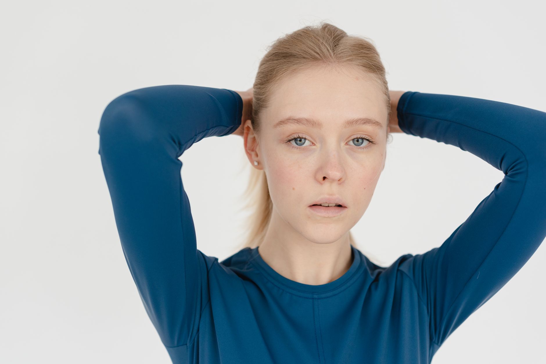Attractive female with blond hair and hands behind head wearing blue wear looking at camera while standing on white background