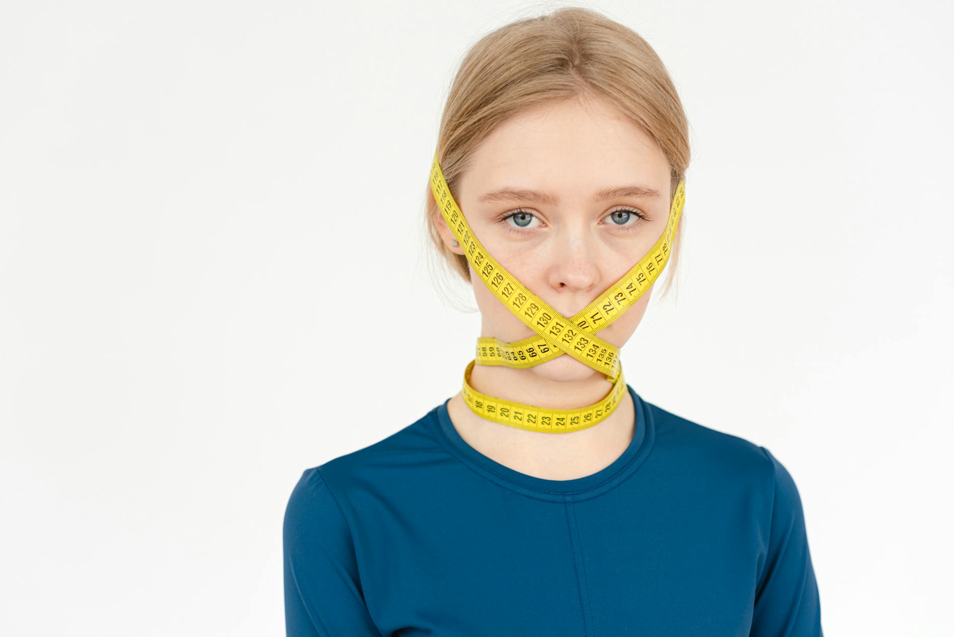 Unhappy young female with centimeter tape on face looking at camera while standing on white background in studio during weight loss