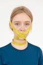 Sad young female with measuring tape on face looking at camera on white background in light studio while losing weight