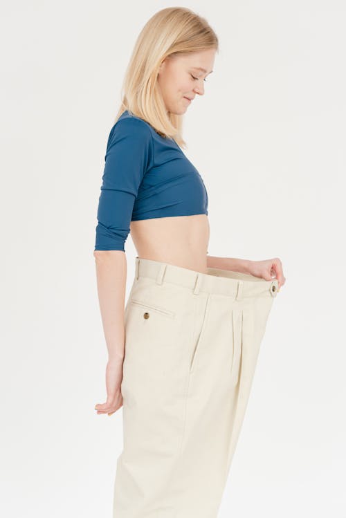 Free Side view of slim positive female with bare belly wearing oversized trousers standing on white background in light studio after weight loss Stock Photo