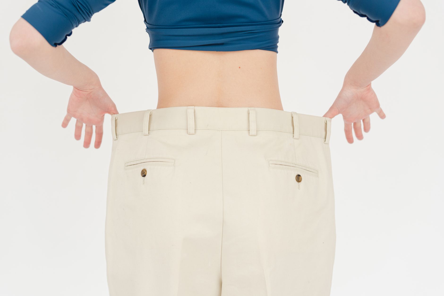 Back view of faceless fit female wearing white oversized trousers standing on white background in light studio after weight loss