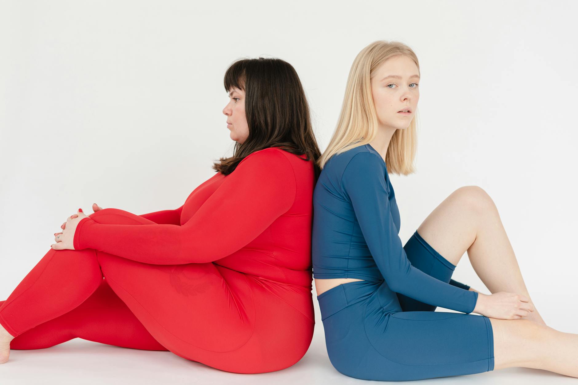 Side view of unhappy plus size female and slender woman in sportswear sitting back to back on white background in studio
