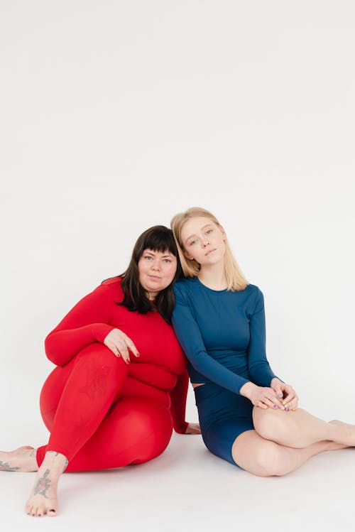 Content plus size barefoot woman and slender female wearing activewear looking at camera while sitting on white background in studio