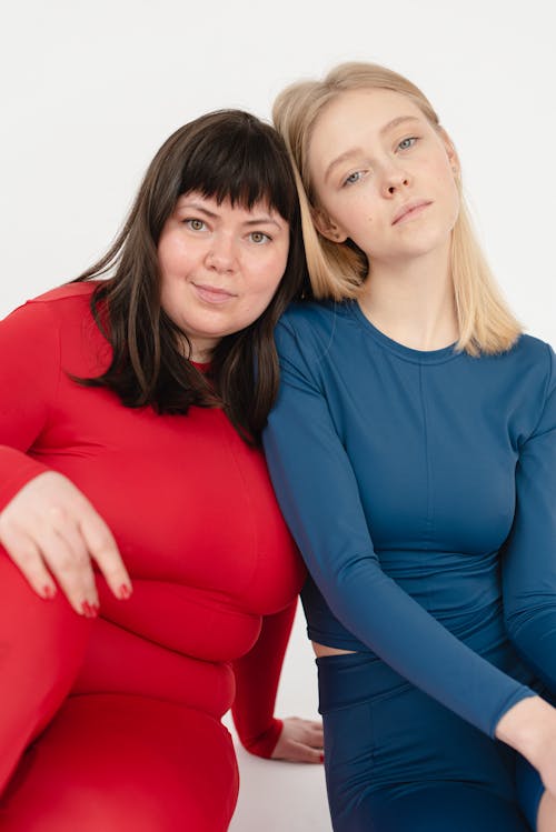Content plump female and slender sportive woman in activewear looking at camera while sitting on white background in light studio