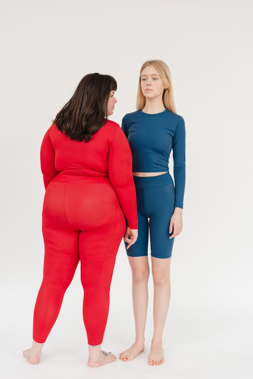 Full body of plus size female in sportswear standing near slim female personal instructor on white background in light studio