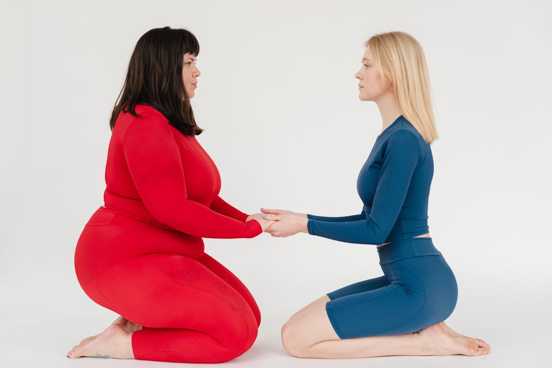 Free Full body side view of plus size woman and slender female instructor in activewear holding hands and looking at each other on white background Stock Photo
