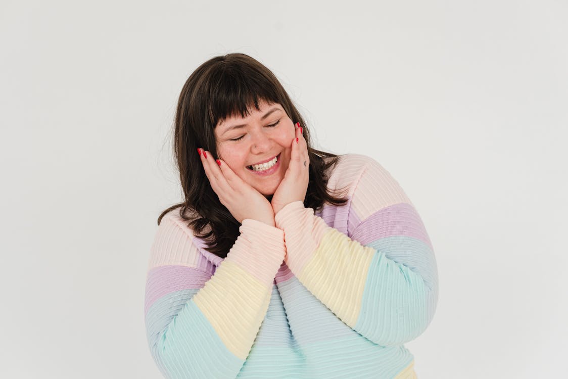 Positive plus size female with closed eyes smiling and touching cheeks while standing on white background in modern light studio