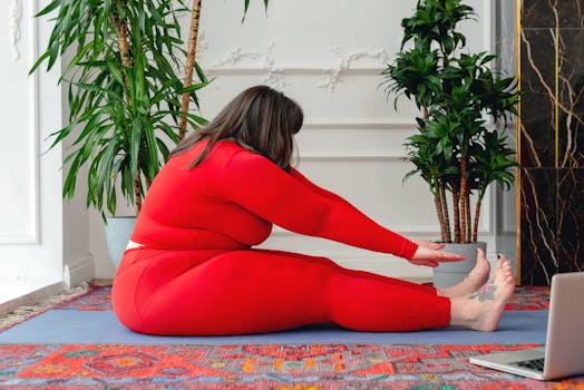 Side view full body of plus size female in red activewear practicing Pascimottanasana posture during online yoga lesson on laptop at home
