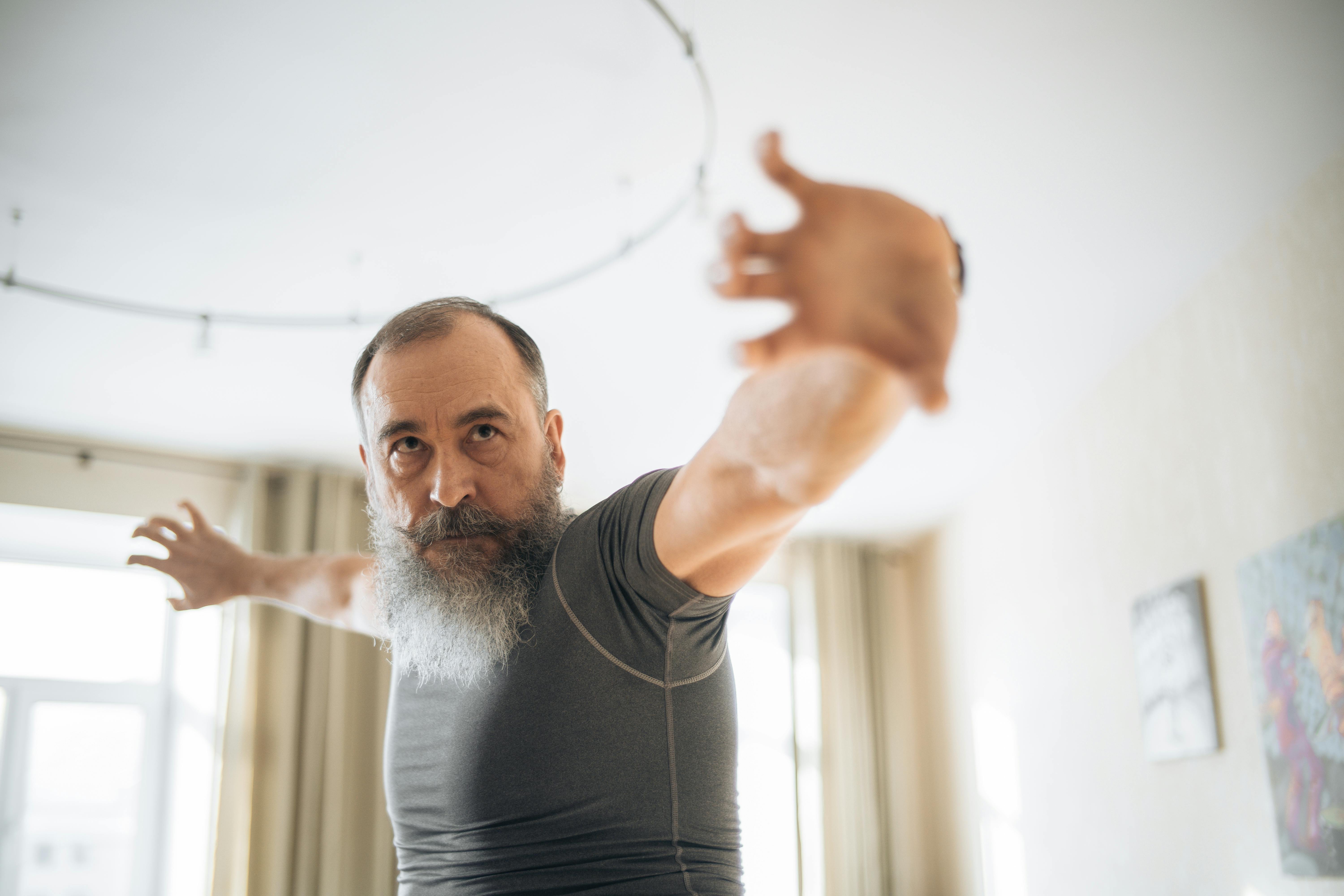 Elderly man with beard doing yoga stretches in a room, enhancing fitness and relaxation.