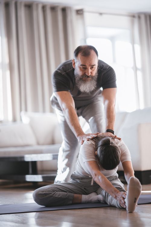 Free Senior man helps with partner stretch in bright indoor setting, promoting flexibility. Stock Photo