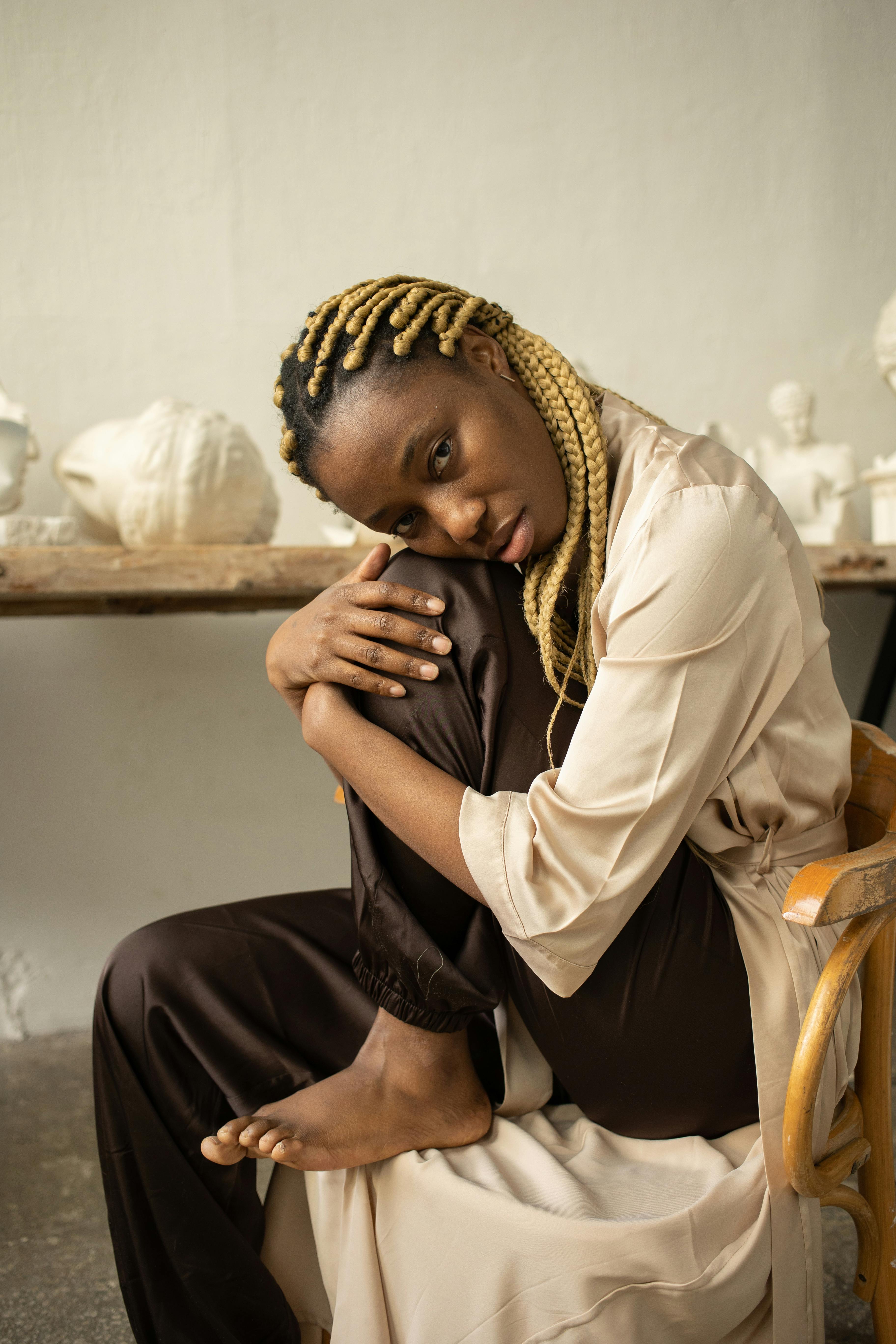 Thoughtful woman with afro braids poses indoors among artistic sculptures.