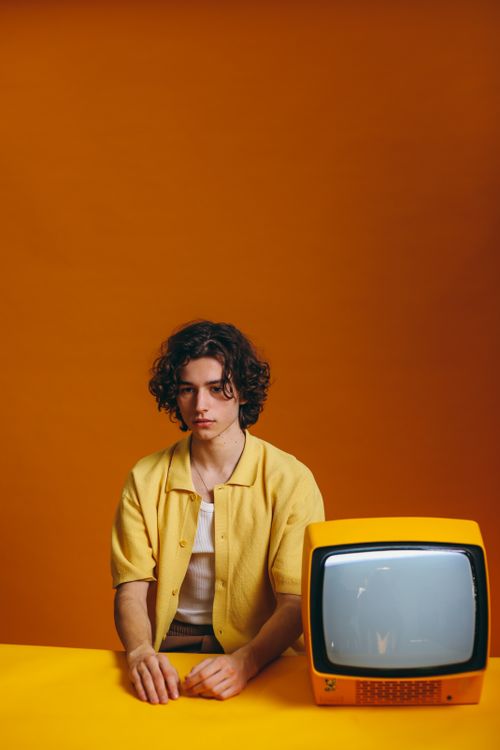Free Young man in yellow attire beside a retro television, orange backdrop. Stock Photo