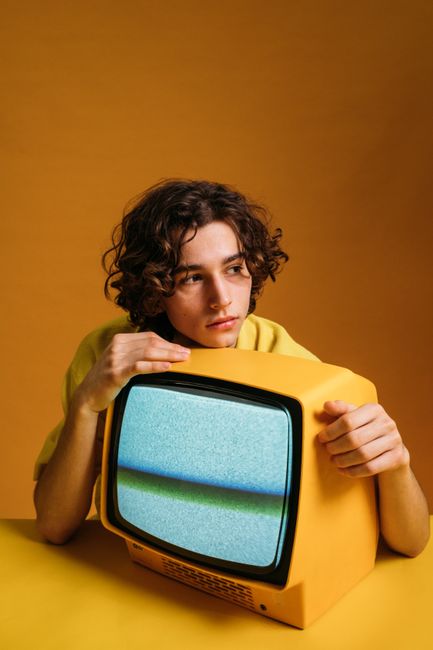Stylish young man with curly hair poses with a vintage yellow TV in a retro setting.