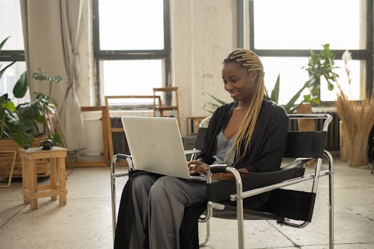 African American woman sitting comfortably using a laptop, working remotely in a cozy indoor space.