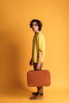 Young man in vintage fashion holding a suitcase against an orange background, exuding retro style.