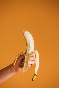 A peeled banana held in hand against a vibrant orange background highlighting its freshness.