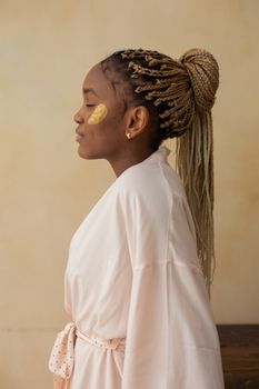 Side view of a woman in a bathrobe applying skincare indoors, showcasing braided hair and relaxation.