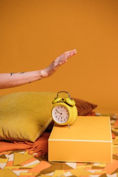 Stylish vintage yellow alarm clock on a colorful background with a woman's hand reaching for it.