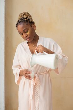 African American woman in bathrobe pouring milk from a pitcher, signifying a relaxed morning routine.