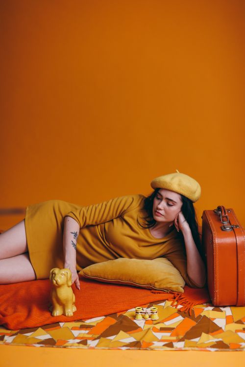 Free Stylish woman in yellow beret and dress poses with orange suitcase in retro-styled room. Stock Photo