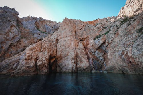 Calm river flowing near rough rocky cliffs with uneven surface against cloudless blue sky in nature on sunny summer day