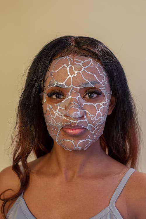 Young calm ethnic female in casual top wearing hydrating facial cloth mask and looking at camera