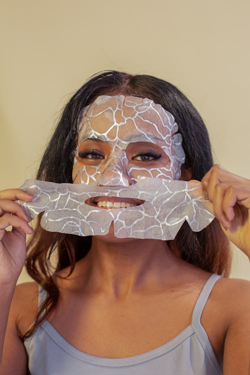 Smiling attractive ethnic female in casual top removing hydrating facial cotton mask and looking at camera against beige wall