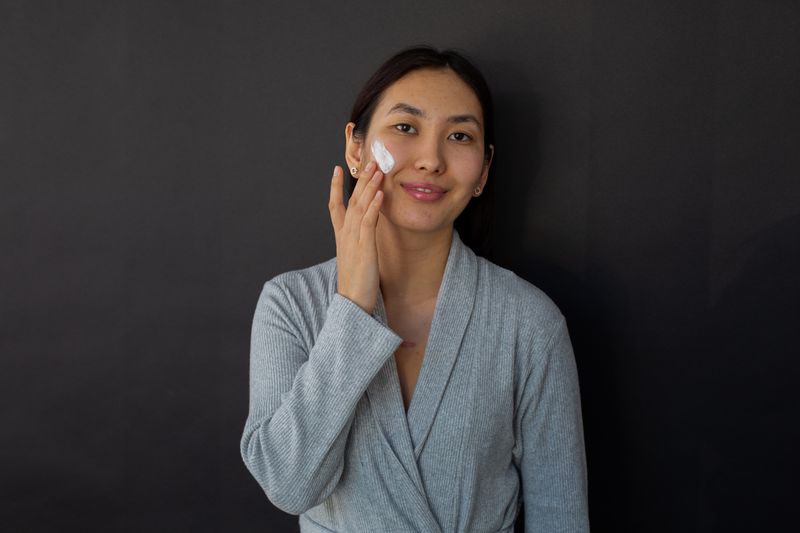 Positive ethnic female in bathrobe applying moisturizing cream on cheek during daily beauty routine against black background and looking at camera