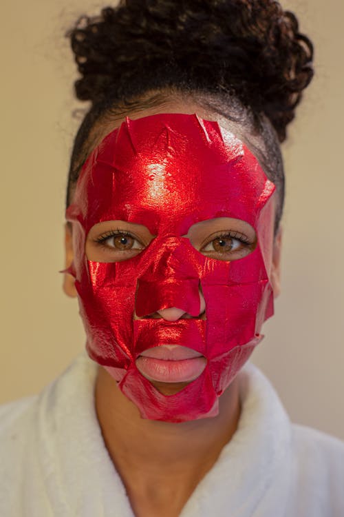 Young African American female in bathrobe doing skin care procedure with bright facial mask and looking at camera against beige background
