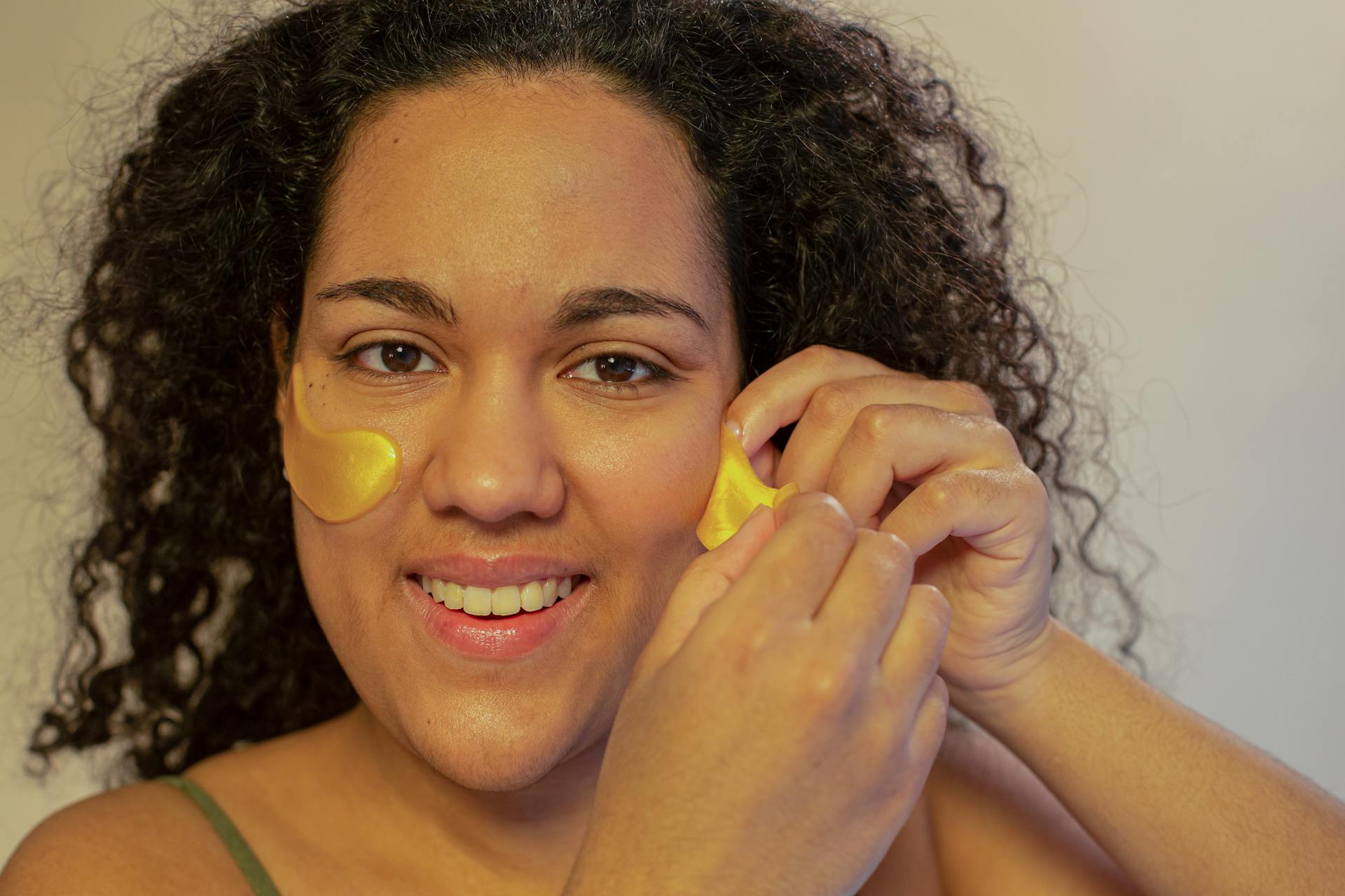 Optimistic ethnic female doing skin care procedure with gel eye patches and looking at camera with toothy smile