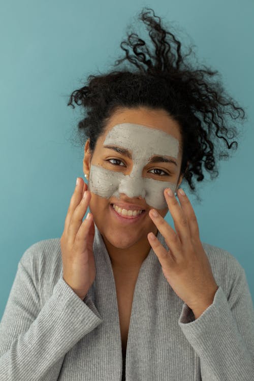 Positive ethnic female applying skincare clay mask for purifying pores and looking at camera against blue background
