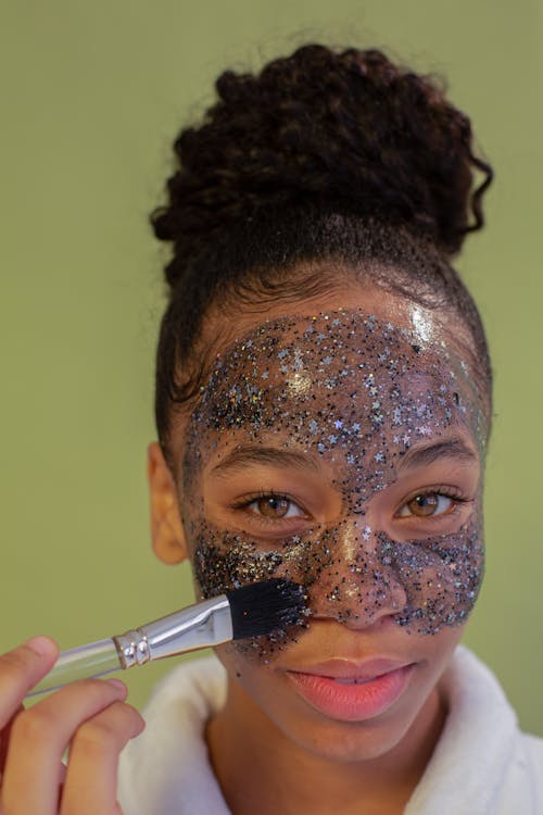 African American female taking care of skin while applying cleansing face mask with brush and looking at camera against green background