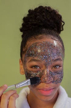 African American female taking care of skin while applying cleansing face mask with brush and looking at camera against green background