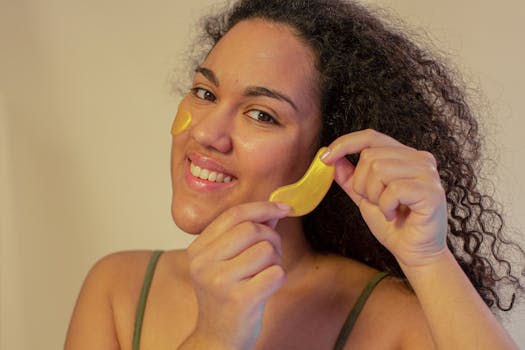 Smiling young woman applying hydrating gold eye patches for skincare routine indoors.