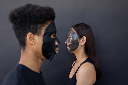 Free Side view of multiethnic couple in sheet masks for cleansing beauty procedure against black background Stock Photo