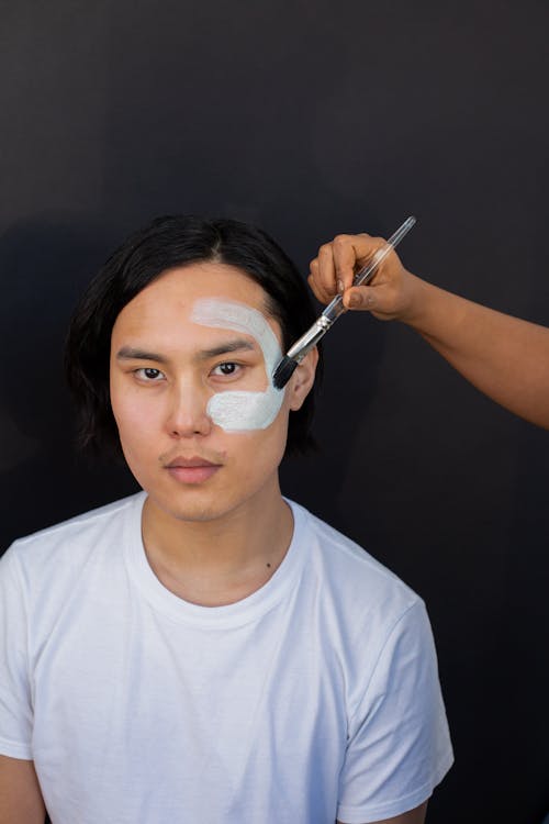 Ethnic cosmetologist applying mask on face of Asian man