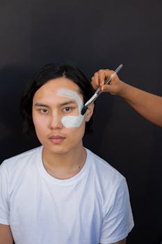 Asian man having a clay mask applied with a brush on a black background.