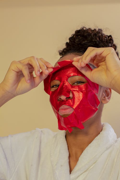 African American female pouting lips while taking off sheet mask on beige background and looking at camera