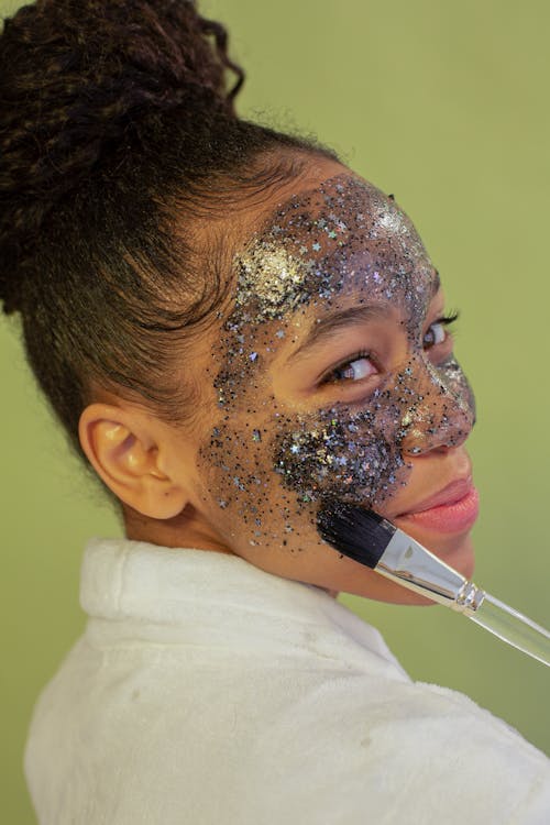 Side view of content ethnic female teenager with brush applying peel off mask with glitter on face while looking at camera on green background