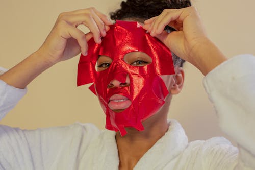 Young ethnic female in bathrobe applying bright hydrating sheet mask on face while looking at camera on beige background